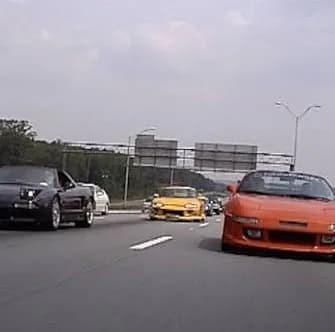 A line of colorful modified 1990s Japanese sports cars driving together on a wide interstate highway during golden hour sunset.
