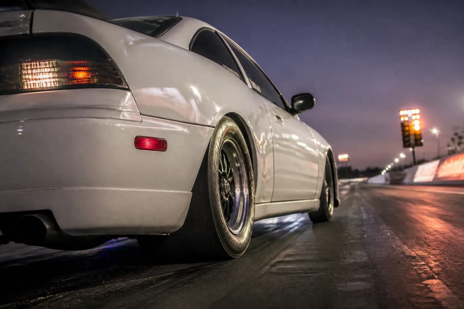 A performance sports car launching off the starting line of a drag strip, showing rear tire squat and dark sticky VHT track prep under stadium lights.