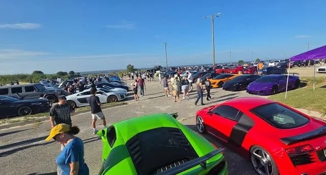 Crowds of car enthusiasts walking among parked exotic supercars including Lamborghinis and an Audi R8 at a sunny outdoor meet.