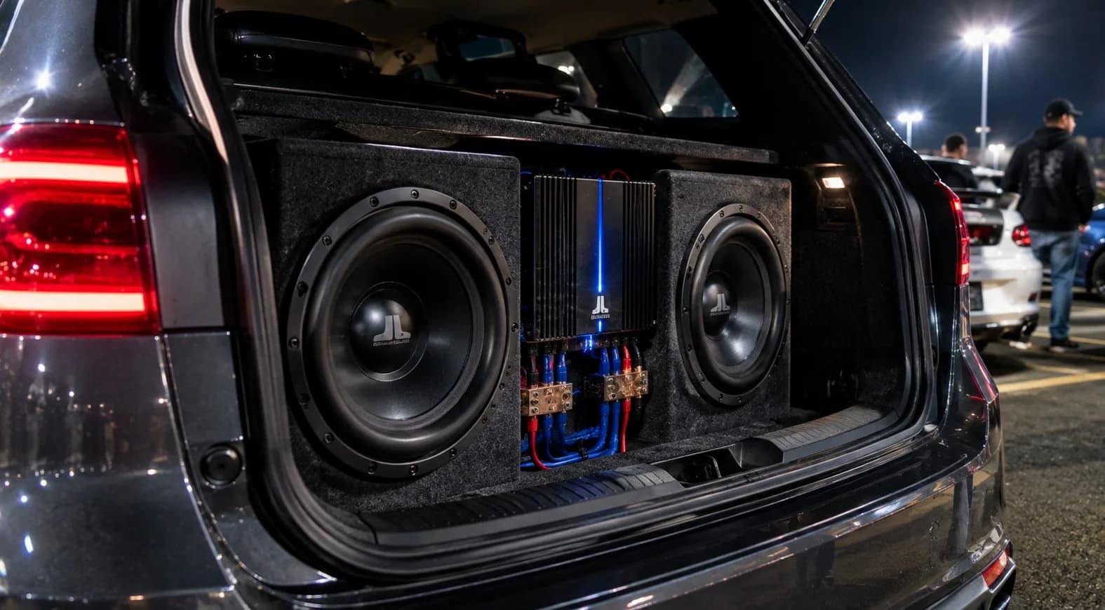 A custom trunk-mounted subwoofer enclosure with dual 12-inch subwoofers, heavy power wiring, and a chrome amplifier illuminated inside a modified car trunk.